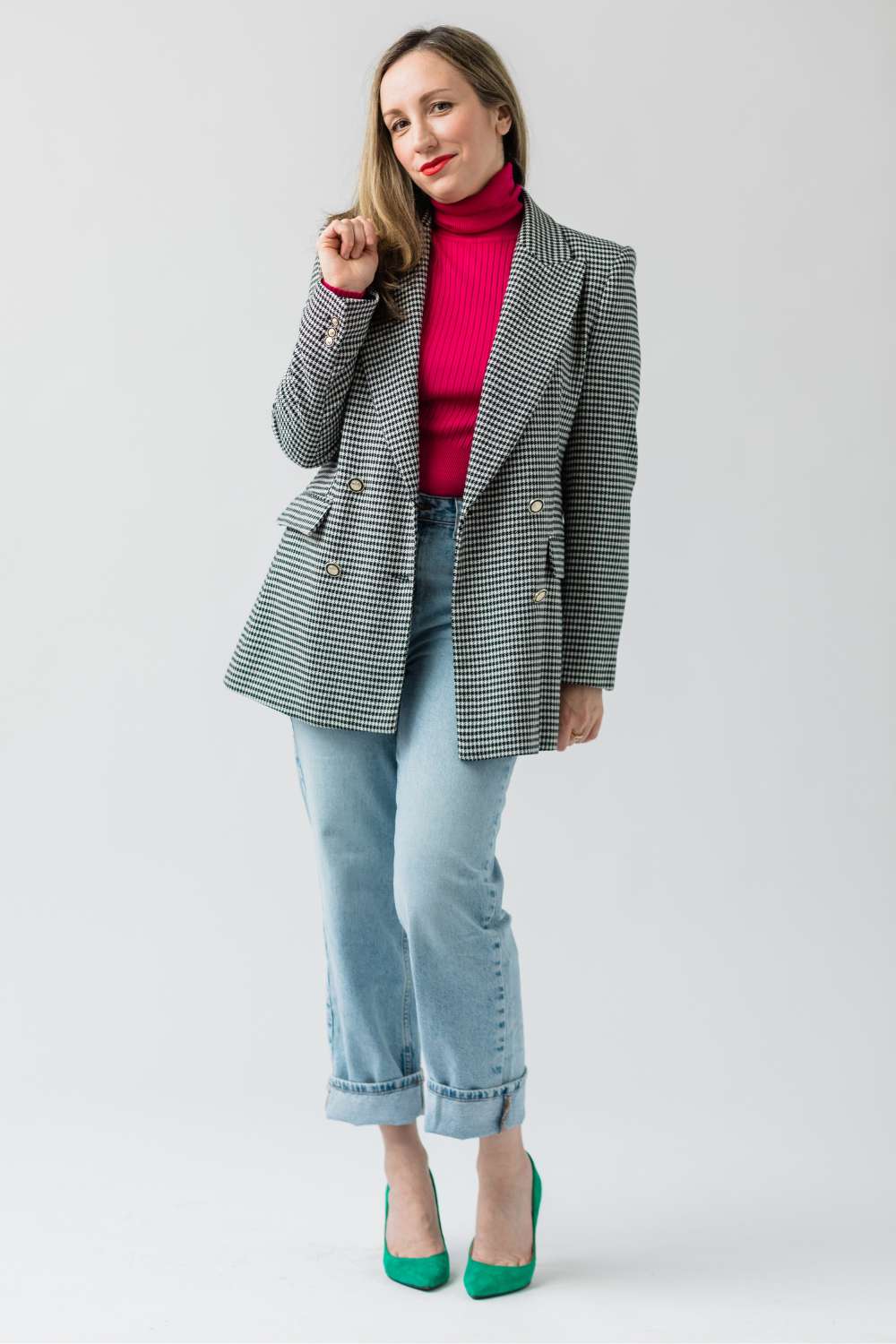 Woman standing and posing wearing a hot pink tshirt, herringbone blazer, light blue jean and green high heel pumps.