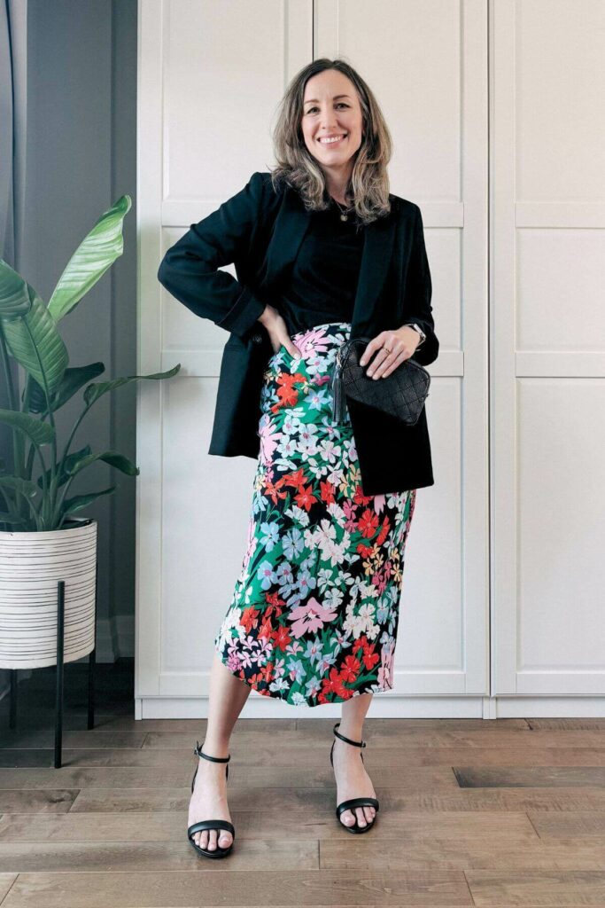 Woman standing in front of white closet wearing a colorful floral midi skirt, black blazer, tshirt and sandals.