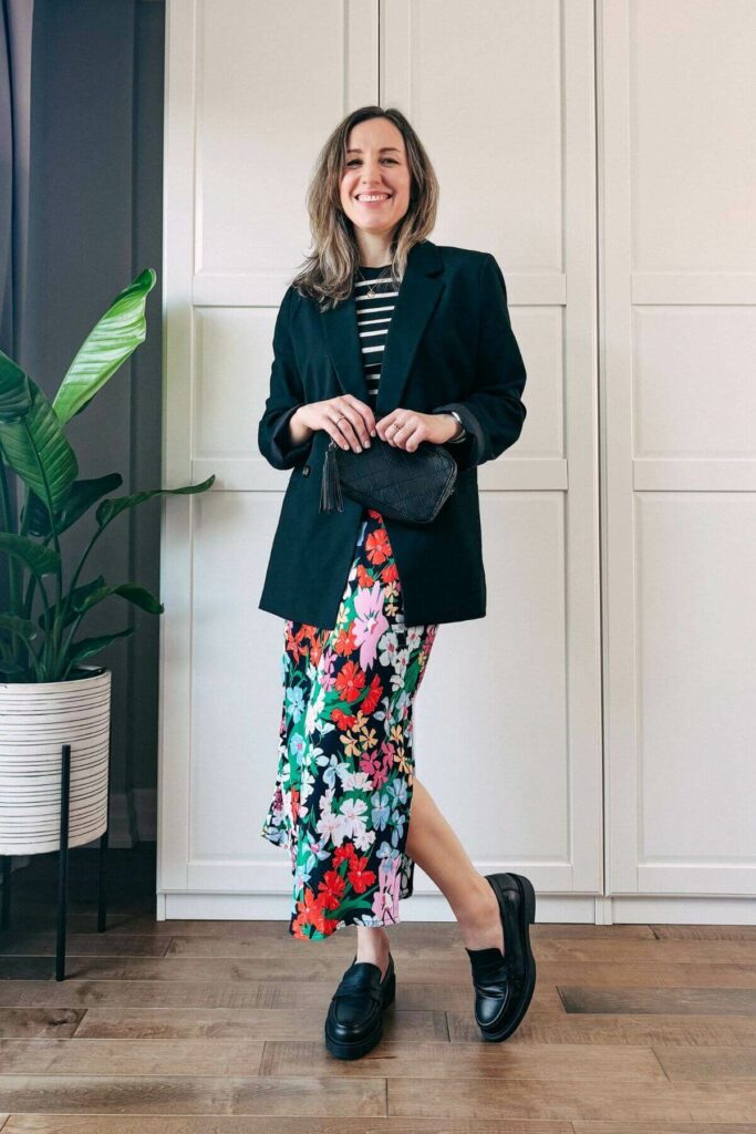 Woman standing in front of white closet wearing a colorful floral midi skirt, black blazer, black loafers and black white striped tshirt.
