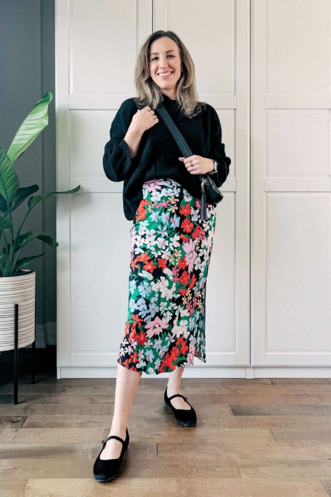 Woman standing in front of white closet wearing a colorful floral midi skirt, black sweater, ballerina flat and purse.