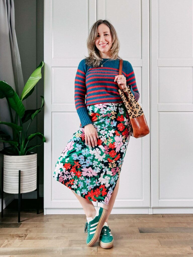 Woman standing in front of white closet wearing a colorful floral midi skirt, striped blue red sweater, leopard print purse, green sneakers.