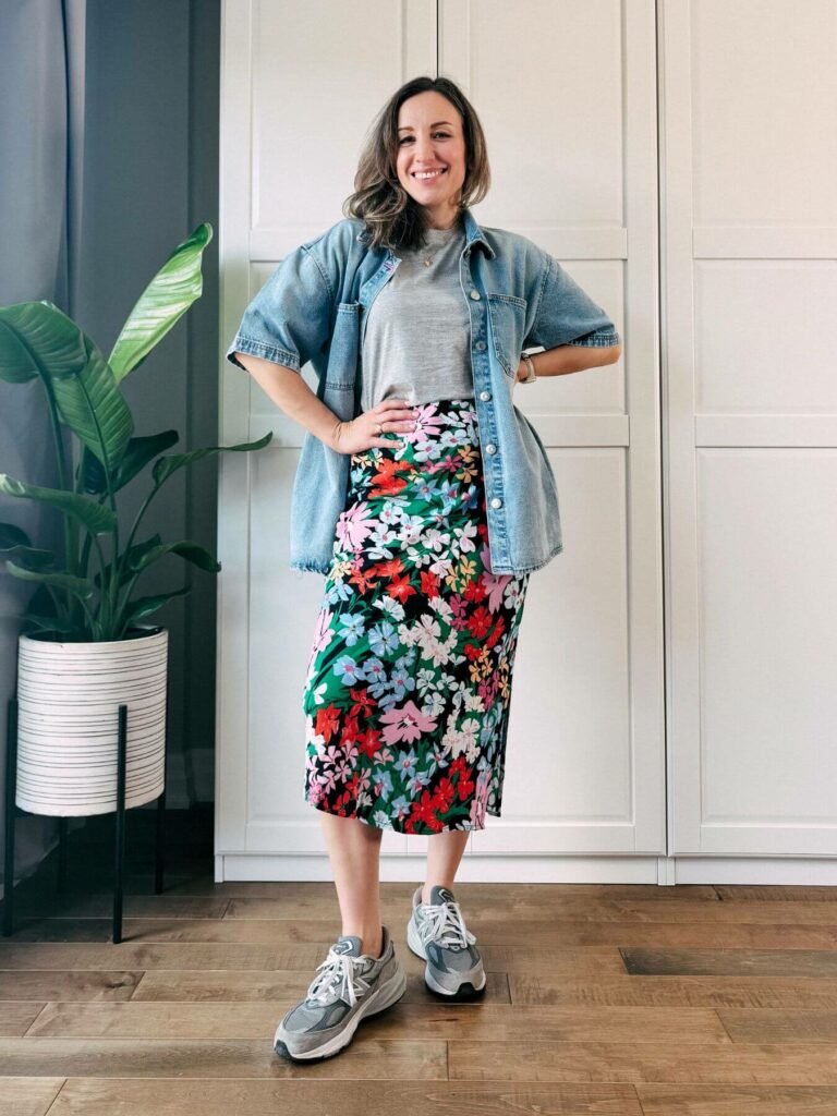 Woman standing in front of white closet wearing a colorful floral midi skirt, grey tshirt, oversized denim shirt, grey athletic sneakers.