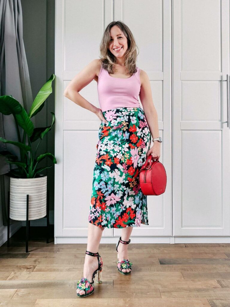 Woman standing in front of white closet wearing a colorful floral midi skirt, pink tank top, floral heels and round red purse.