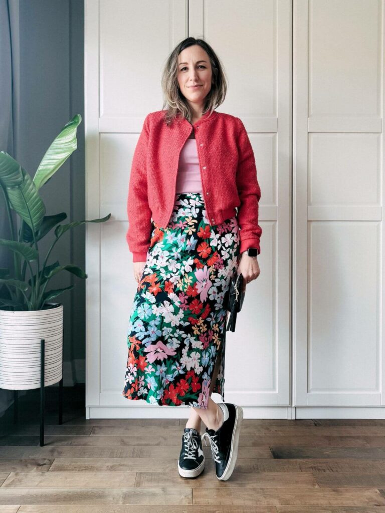 Woman standing in front of white closet wearing a colorful floral midi skirt, pink tank top, red cropped jacket and black whit sneakers.