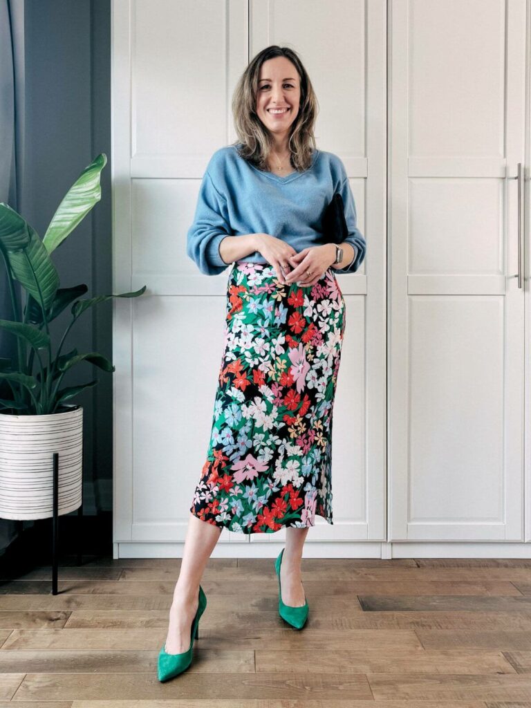 Woman standing in front of white closet wearing a colorful floral midi skirt, light blue sweater and green high heels.