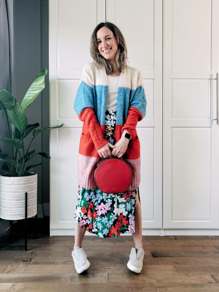 Woman standing in front of white closet wearing a colorful floral midi skirt, beige sneakers, colorful cardigan and round red purse. 