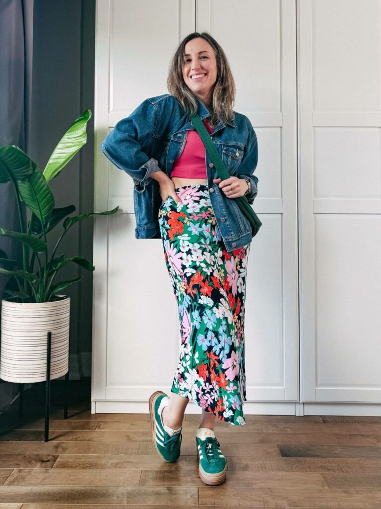 Woman standing in front of white closet wearing a colorful floral midi skirt, hot pink tshirt, oversized denim jacket, green sneakers and crossbody bag.