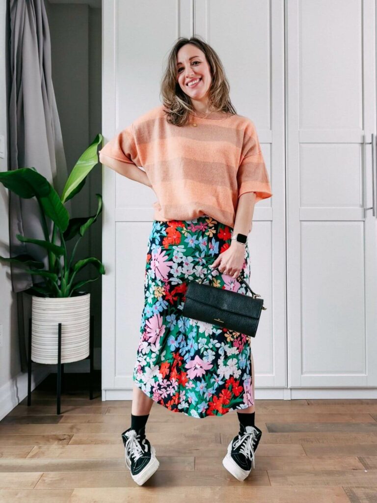 Woman standing in front of white closet wearing a colorful floral midi skirt, orange striped sweater, black high top sneaker and black purse.