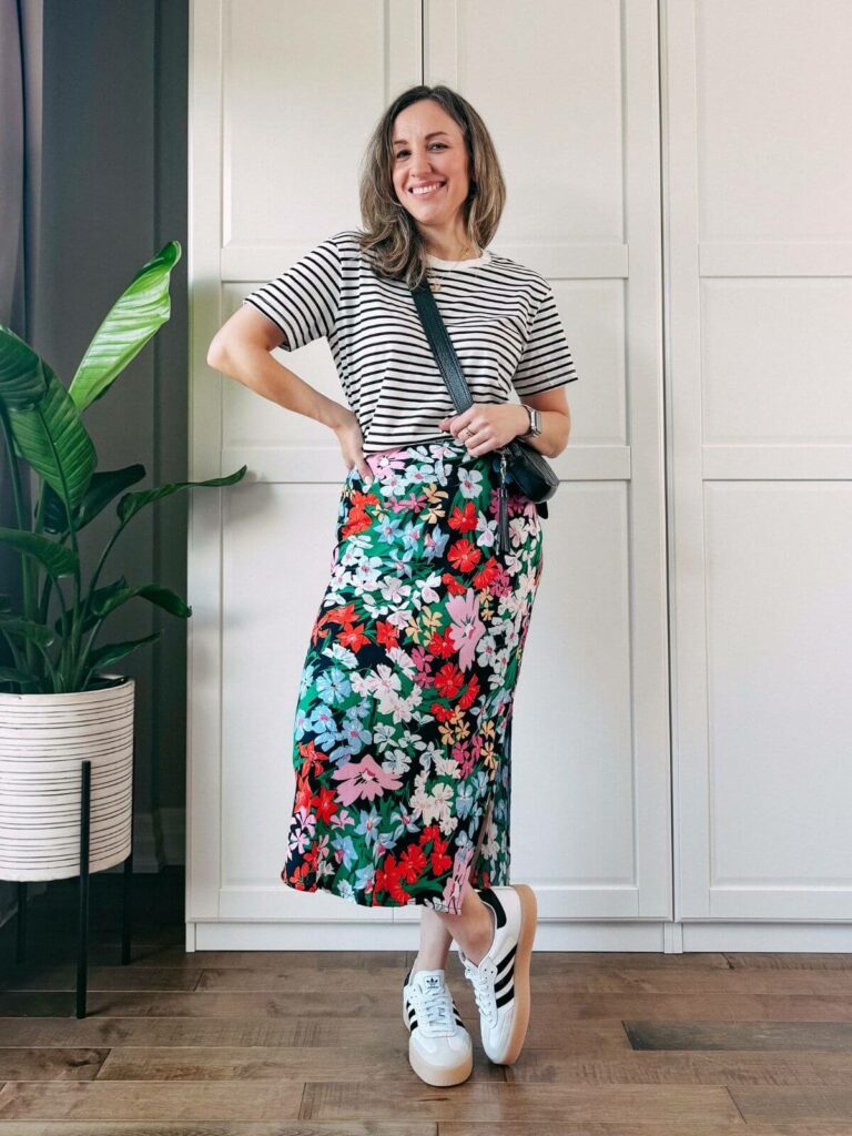 Woman standing in front of white closet wearing a colorful floral midi skirt, black white striped tshirt, white sneakers and black purse.