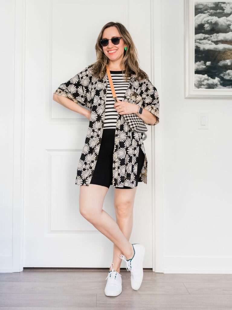 Brunette woman posing in a summer outfit wearing white and black printed tank and shirt, black bike shorts and white knit sneakers.