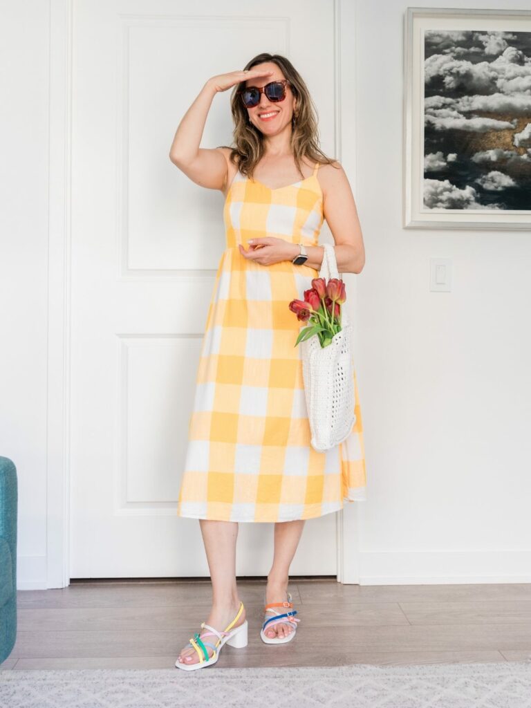 Brunette woman posing in a summer outfit wearing a white and yellow large print gingham midi dress, colorful heeled sandals and carrying a white crochet tote with tulips inside.