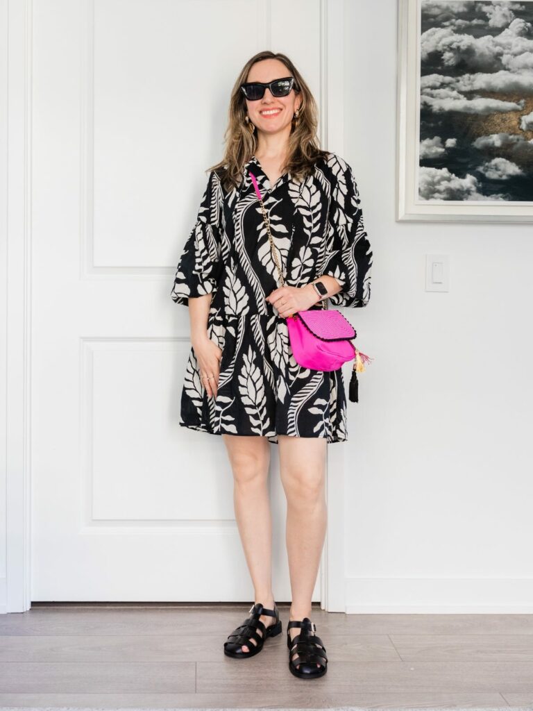 Brunette woman posing in a summer outfit wearing white and black printed mini dress, hot pink purse and black fisherman sandals.