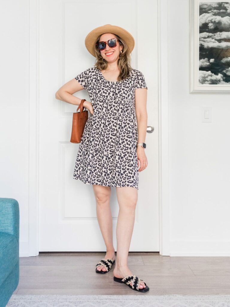 Brunette woman posing in a summer outfit wearing leopard print mini dress and sandals with wide brim hat.