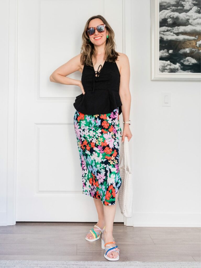 Brunette woman posing in a summer outfit wearing colorful floral midi skirt, black peplum halter and sandals.