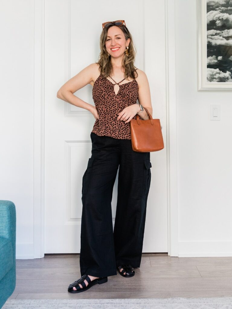 Woman posing wearing a brown cheetah print camisole top, black linen pants and black fisherman sandals holding a brown handbag.