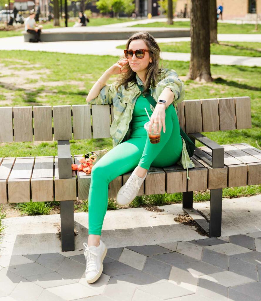 Brunette lady sitting on a wooden park bench wearing bright green leggings and tank with a light camo green jacket and off white sneakers.  