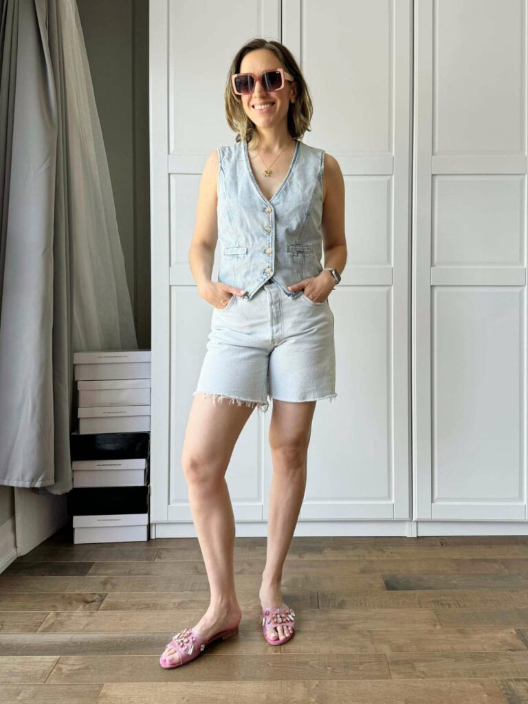 Woman posing in front of a white closet wearing a light denim vest and matching light denim shorts and pink jewelled slide sandals and brown sunglasses.