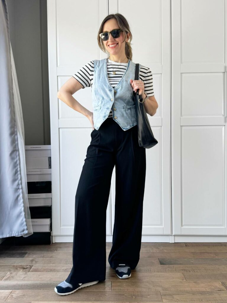 Woman posing in front of a white closet wearing a light denim vest styled with black wide legs pants and striped T-shirt.