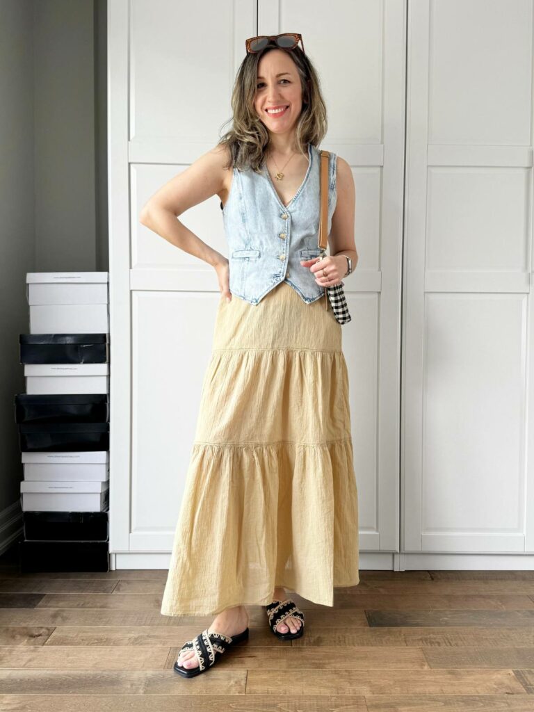 Woman posing in front of a white closet wearing a light denim vest styled with long wide light camel skirt.