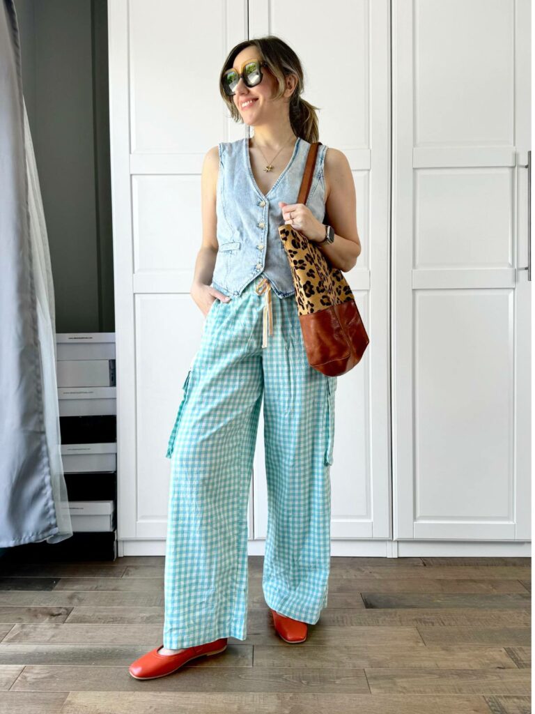 Woman posing in front of a white closet wearing a light denim vest styled with checked wide legs pants.