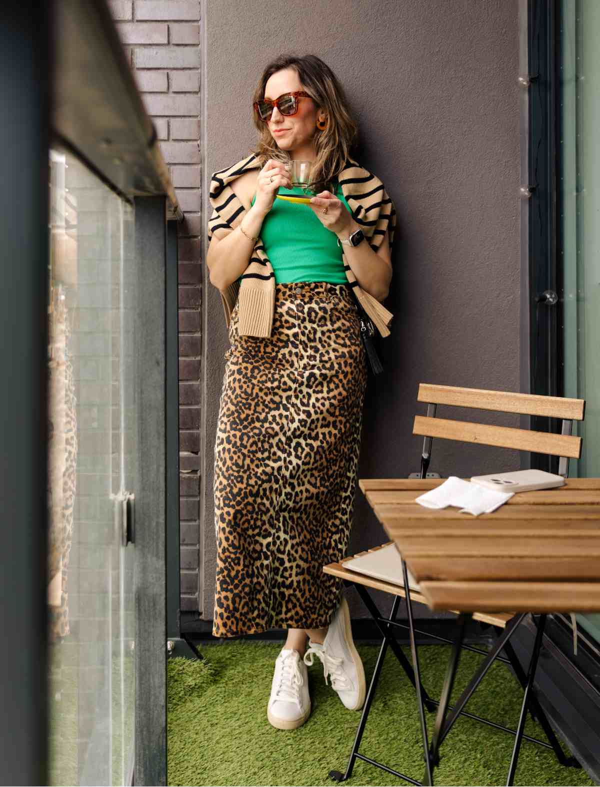 Woman posing on balcony wearing a leopard print skirt, green tank top, tan and black striped sweater and white sneakers.