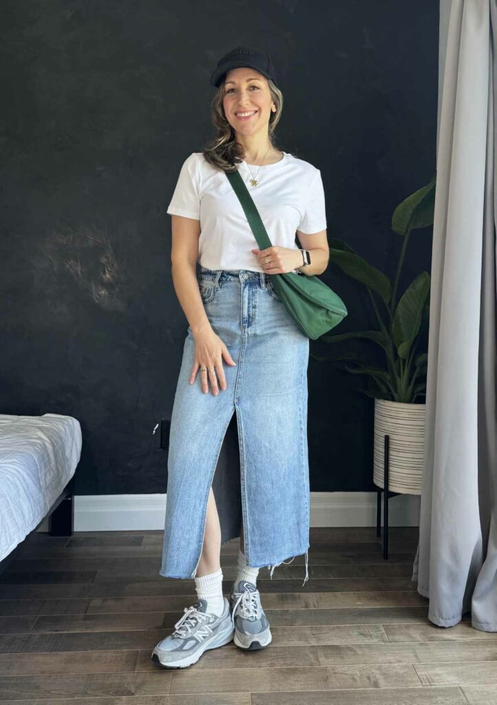 Woman posing in front of black wall wearing a long denim skirt with basic white tee, green crossbody bag and chunky sneakers.