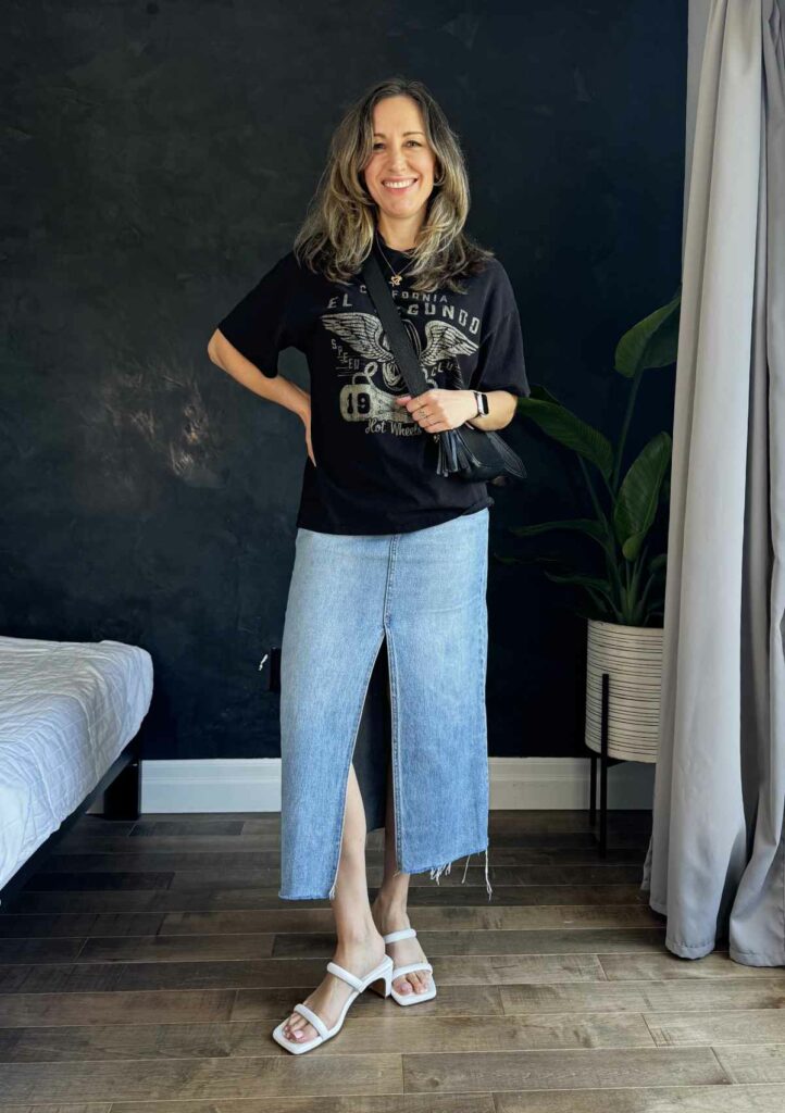 Woman posing in from of black wall wearing a long denim skirt, black graphic tee and white sandals.