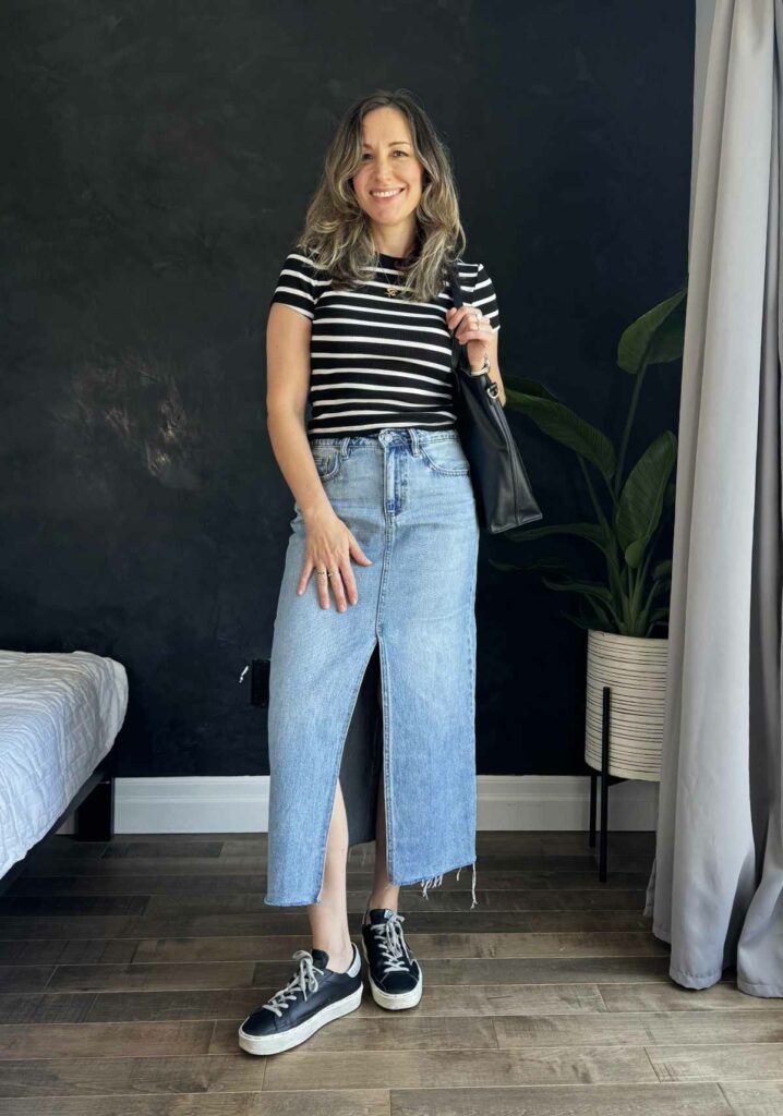 Woman posing in from of black wall wearing a long denim skirt, striped tee and black sneakers.