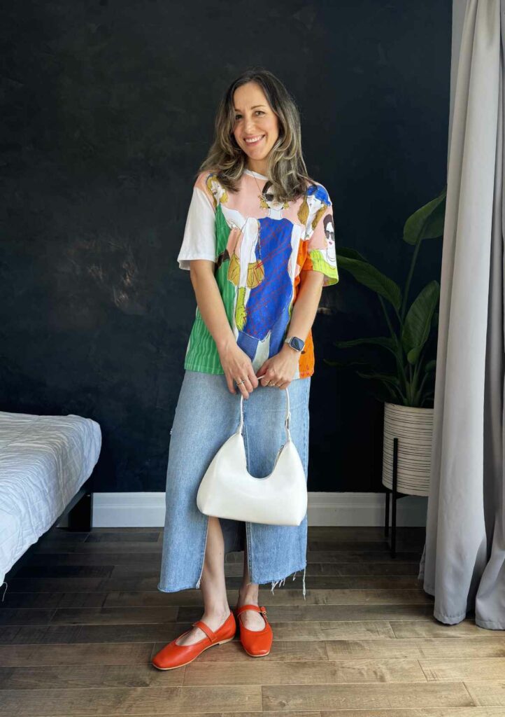 Woman posing in from of black wall wearing a long denim skirt, graphic t-shirt and red ballet flats.