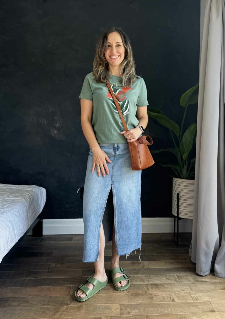 Woman posing in from of black wall wearing a long denim skirt, graphic green tshirt and green sandals.