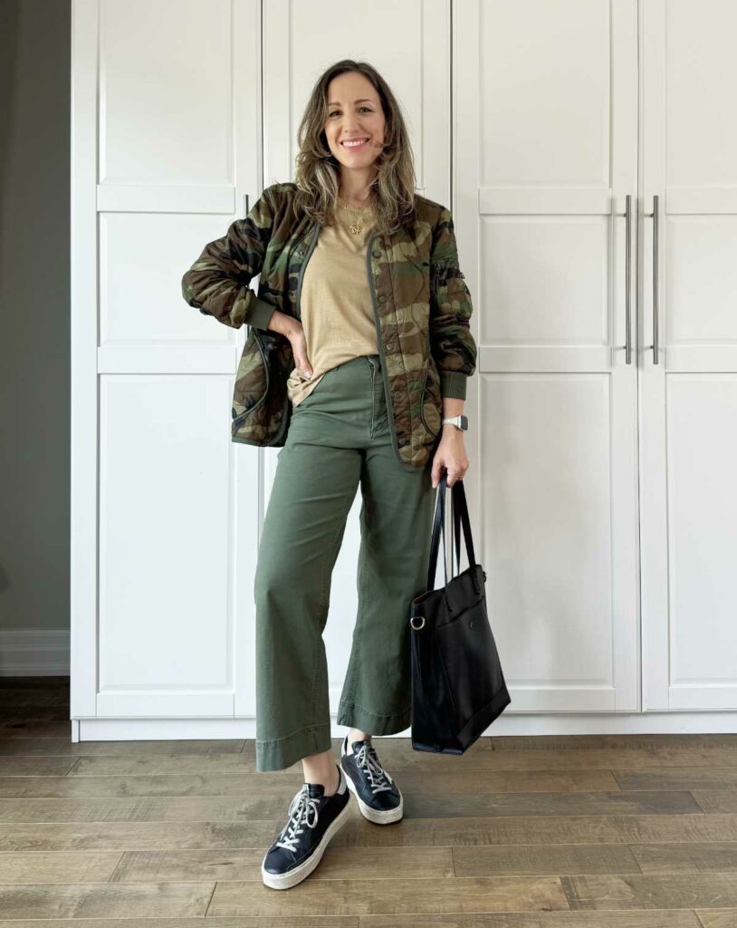 Woman posing wearing olive green pants for fall with dark beige tee, camo jacket + sneakers.