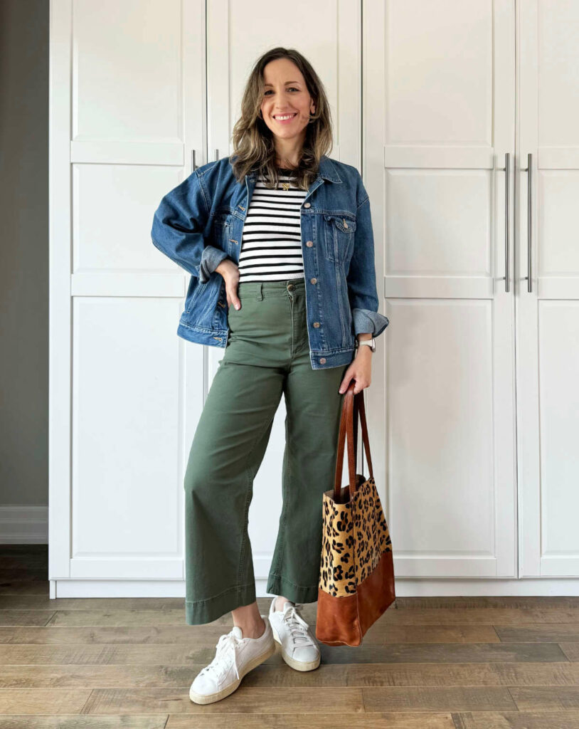 Woman posing wearing olive green pants for fall with denim jacket, striped top + leopard print tote.